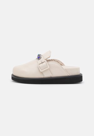 Beige lædersandal med sort sål, der har en dekorativ metalspænde og blødt indre for komfort.