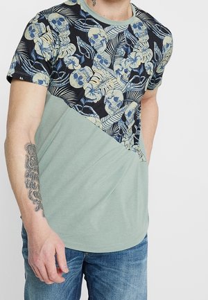 Chemise à manches courtes avec une partie supérieure foncée à motifs de crânes et de feuillage, et une section inférieure unie verte. Matière en coton doux.