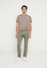 Καφέ κοντομάνικο μπλουζάκι με ένα μικρό γραφικό λογότυπο, συνδυασμένο με εφαρμοστά ελιάς πράσινα παντελόνια και λευκά αθλητικά παπούτσια. Λείες υφές, άνετη εφαρμογή.