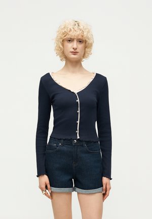 Osoba s kučeravými blond vlasmi oblečená v tmavomodrej zapínanej blúzke s dlhým rukávom a vyhrnutých tmavých rifľových kraťasoch na jednotnom pozadí.