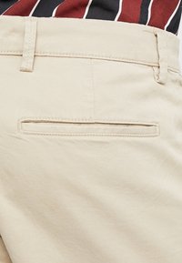 Beige katoenen broek met een horizontale paspelzak aan de achterkant, met een gladde textuur en een taillering.