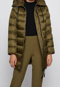 Olivově zelená prošívaná bunda s kapucí, s horizontálním prošíváním, předním zipem a černými šňůrkami. Kombinována s úzkými kalhotami.