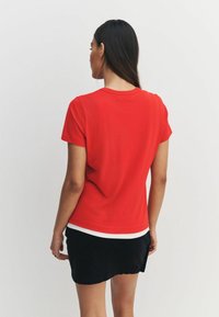 Sarkans īsroku T-krekls ar apaļu apkakli, ar baltu uzsvaru apakšmalā, izgatavots no gluda kokvilnas materiāla. Redzams no aizmugures.