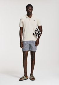 Krém színű póló, csíkozott fürdőnadrág és barna szandál. Fekete-fehér spirálmintás törölközőt tart. Simaságú szövettextúrák.