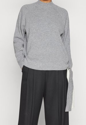 Personne portant un pull en maille gris avec des manches longues et des pantalons larges noirs, les mains partiellement dans les poches, sur un fond uni.