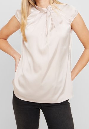 Femme portant un chemisier en satin de couleur crème à manches courtes avec un décolleté en trou de serrure et un pantalon noir, se tenant les mains sur les hanches.