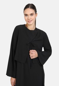 Schwarze, kurz geschnittene Jacke mit langen, ausgestellten Ärmeln, aus texturiertem Stoff, mit einer Frontbindeschließe und getragen über einem passenden schwarzen Kleid.