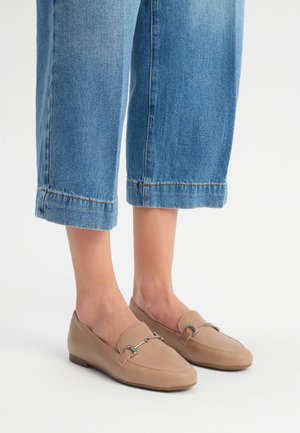 Mocassini beige in pelle liscia, con un accento di catena argentata e punta rotonda. Indossati con pantaloni in denim blu a vita corta.