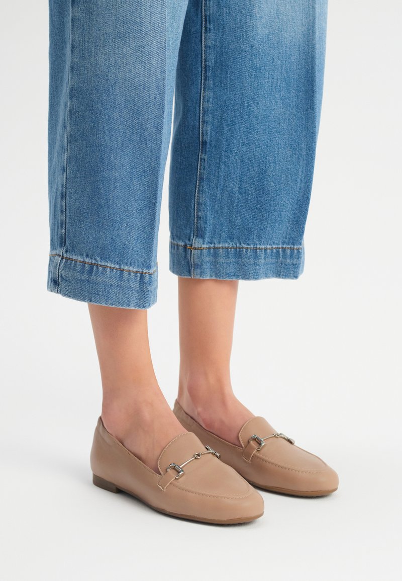 Beige loaferit, valmistettu sileästä nahasta, jossa on hopeinen ketjuyksityiskohta ja pyöristetty kärki. Yhdistettynä lyhyisiin sinisiin farkkuihin.