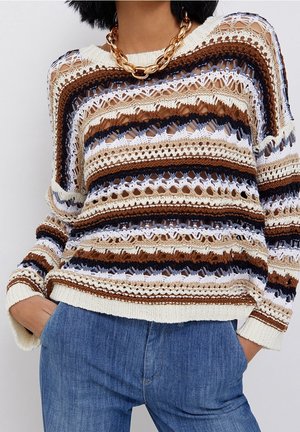 Vrouw draagt een gestreepte opengewerkte trui in crème, bruin, zwart en blauw, met een blauwe spijkerbroek en een dikke gouden ketting.