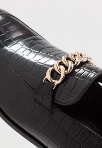 Mocassin noir en cuir synthétique texturé avec un détail en chaîne sur le dessus, présentant une finition brillante, une semelle plate et des accents cousus.