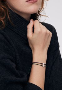 Skórzana bransoletka z dwoma perłami i małymi metalowymi akcentami, z wieloma cienkimi sznurkami w kolorze czarnym i zielonym na tle ciemnego swetra.