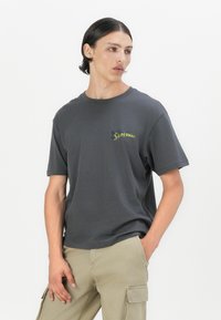 Camiseta de algodón gris de manga corta, con un colorido logo de "Superman" en el lado izquierdo del pecho. Corte casual, textura suave, diseño de cuello redondo.
