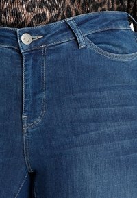 Acercamiento a la cinturilla de unos pantalones de mezclilla azul con botón de metal, presillas para el cinturón y costuras del bolsillo delantero.