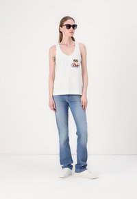 Witte katoenen tanktop met een ronde halslijn, voorzien van een rood en blauw logopatch. Gepaard met lichtblauwe jeans en witte sneakers.