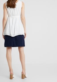 Woman wearing sleeveless white blouse and knee-length navy skirt, standing against plain light gray background, back facing viewer.