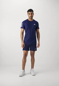 Námornícky modré športové tričko a šortky z priedušnej látky. Na rukávoch sú tri biele pruhy, na hrudi logo a biele tenisky.