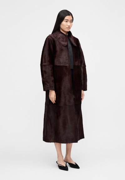 Moteris dėvi ilgą, tamsiai rudą kailinį su plačia apykakle ir juodus aukštakulnius su atviru kulnu, stovinti priešais neutralią foną.