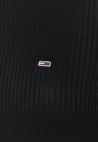 Maglietta in tessuto nero a coste con un piccolo logo ricamato in rosso, bianco e blu. La texture è liscia con strisce verticali.