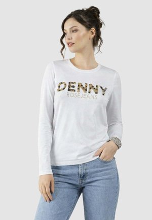 Donna con capelli scuri raccolti in uno chignon, indossa una camicia bianca a maniche lunghe con la scritta "DENNY ROSE JEANS" e jeans azzurri, con la mano in tasca.