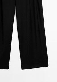 Pantaloni neri a gamba larga mostrati stesi, con la parte inferiore di entrambe le gambe visibile su uno sfondo bianco semplice.