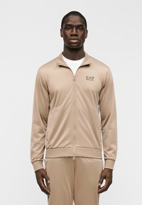 Μπεζ ζακέτα φόρμας από λείο ύφασμα με φερμουάρ μπροστά, όρθιο γιακά και διακριτικό λογότυπο στη αριστερή πλευρά. Διαθέτει μακριά μανίκια.