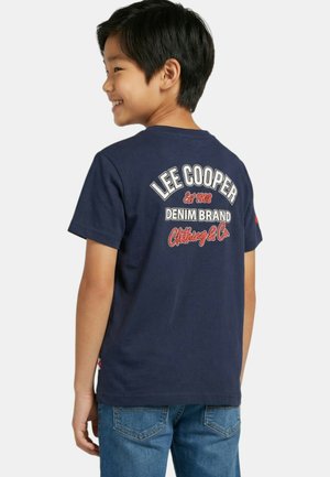 Garçon souriant portant un t-shirt Lee Cooper bleu marine avec le logo de la marque et un texte au dos, assorti à un jean bleu, vu de profil.