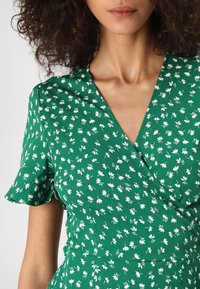 Robe portefeuille verte à motifs floraux, manches courtes, encolure en V plongeante, en tissu léger orné d'un petit motif de fleurs blanches.