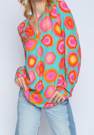 Femme portant un chemisier à manches longues aux motifs vifs avec des hexagones roses, orange et jaunes sur fond turquoise et un jean bleu clair.