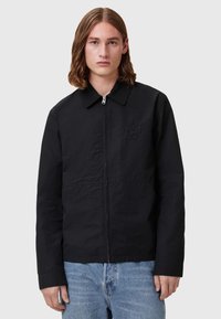 Čierna bunda na zips s golierom, so subtilným embosovaným logom, rovný strih a ľahký materiál. Kombinovaná s svetlomodrými džínsami.