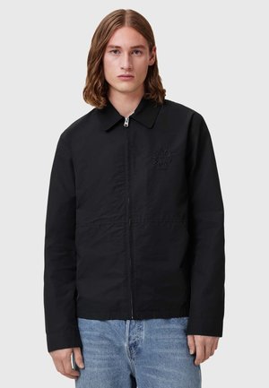 Schwarze Zip-Jacke mit Kragen, mit einem dezenten geprägten Logo, gerader Schnitt und leichtem Material. Getragen mit hellblauen Jeans.