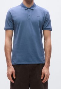 Homme portant un polo bleu à manches courtes avec col et boutons, associé à un pantalon foncé, debout devant un fond uni.