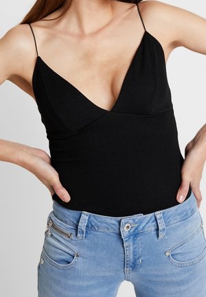 Femme portant un top noir sans manches à col en V avec de fines bretelles et un jean denim bleu clair, mains sur les hanches.