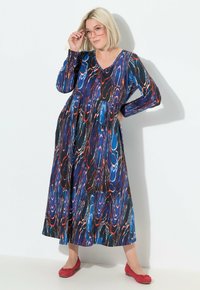 Abito lungo e fluido con scollatura a V e maniche lunghe. Il tessuto presenta un motivo ondulato blu, nero e rosso. Abbinato a ballerine rosse.