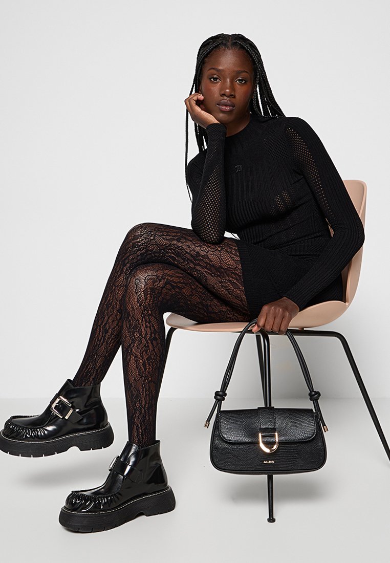 Jeune femme aux cheveux tressés assise sur une chaise portant une robe en maille noire, des collants en dentelle, des chaussures noires épaisses, tenant un sac à main noir par ses anses.