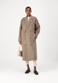 Manteau en laine beige avec un design croisé, de grands revers et des poches avant. Le modèle tient un sac à main rose clair, portant un pantalon clair et des mocassins.