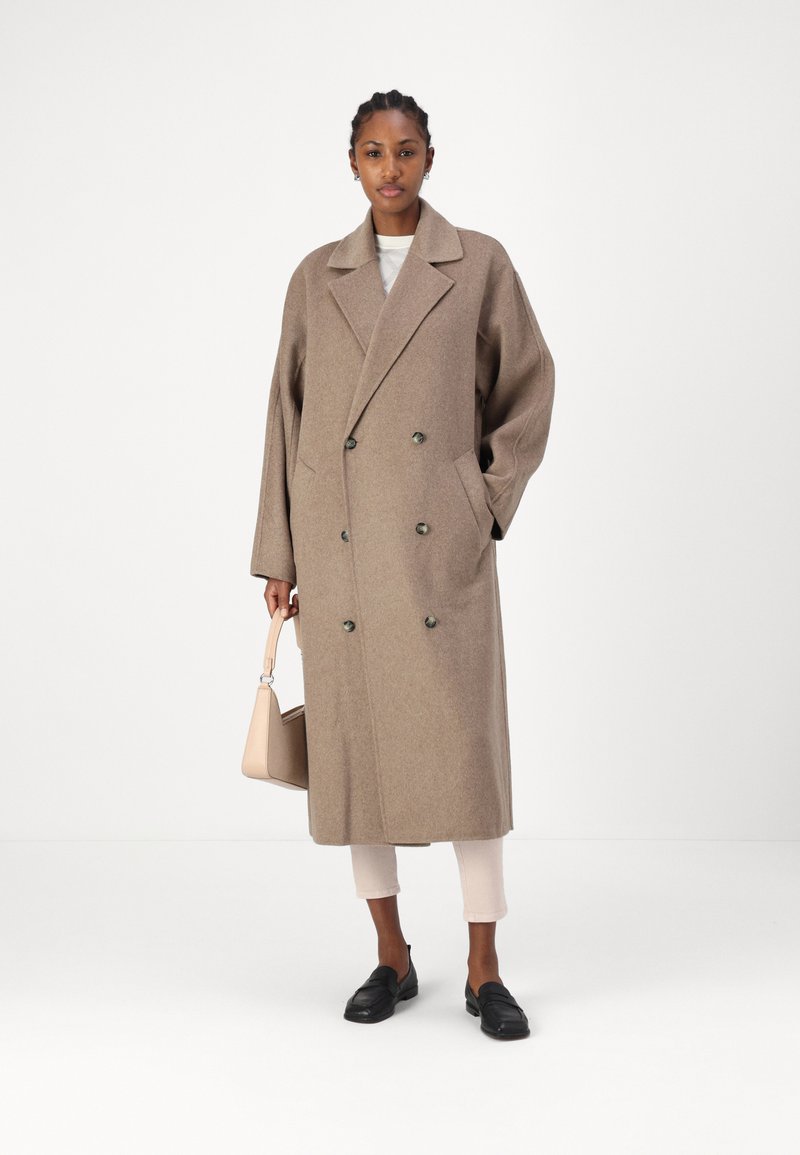 Beige Wollmantel mit doppelreihigem Design, großen Revers und vorderen Taschen. Das Model hält eine hellrosa Handtasche, trägt helle Hosen und Slipper.