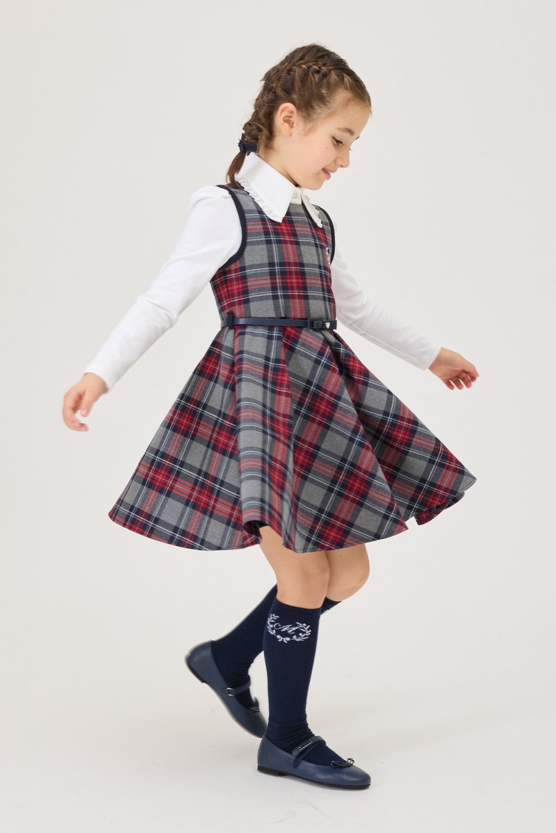Fille tournoyant dans une robe à carreaux rouge et grise avec une chemise blanche, des chaussettes hautes bleu marine et des chaussures Mary Jane bleu marine sur un fond clair.