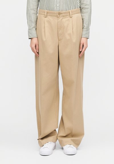 Persoană purtând pantaloni lejeri bej, adidași albi și o cămașă cu dungi verticale cu mâneci lungi, stând în picioare pe un fundal simplu.