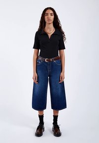 Zwart geribbeld poloshirt, wijde, hooggetailleerde denim culottes, bruine riem met zilveren gesp, donkere sokken en dikke zwarte schoenen.