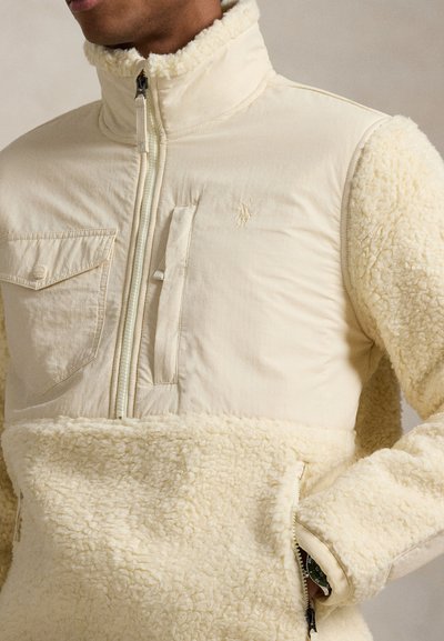 Kreemikas fliisjope kõrge kraega, millel on tõmblukuga sulgemine ja taskud. Pehme tekstuur koos kontrastse siledama ülaosaga.