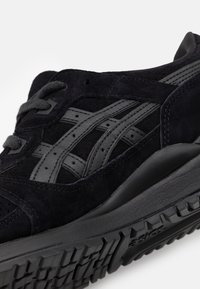 Černá semišová teniska se šedými koženými akcenty, vybavená texturovanou podrážkou a výraznými překříženými proužky po stranách.