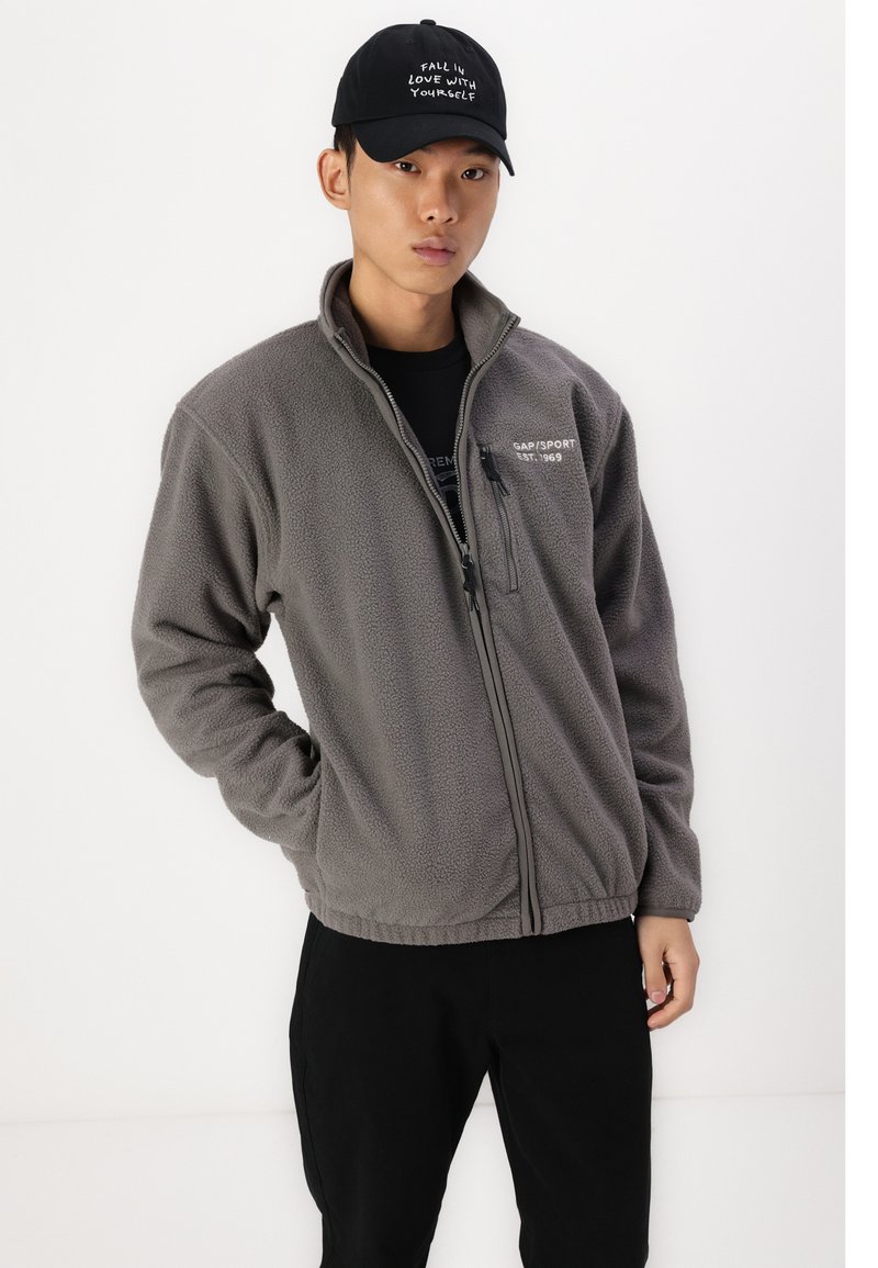 Graue Fleecejacke mit Reißverschluss vorne und seitlichen Taschen. Mit einem Logo auf der Brust und einem strukturierten Stoff. Getragen mit einer schwarzen Mütze.