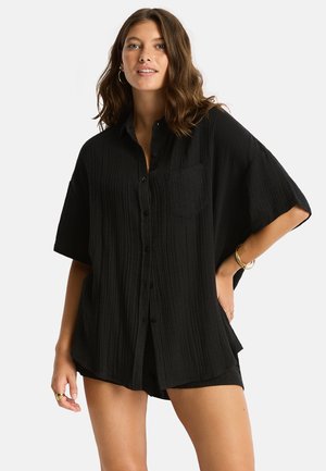 Jeune femme aux cheveux bruns ondulés porte une chemise noire ample boutonnée et un short assorti, accessoirisés de boucles d'oreilles créoles dorées et de bracelets.