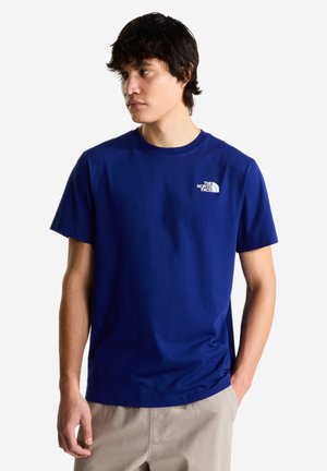 Ung mand iført en blå The North Face t-shirt og beige bukser, stående med den ene hånd i lommen og kigger mod venstre mod en hvid baggrund.