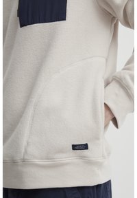 Sudadera de forro polar crema con bolsillo de tela azul marino. Cuenta con costuras laterales, una superficie texturizada y una etiqueta con texto blanco en el dobladillo.