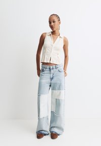 Lichtgekleurde, gestructureerde mouwloze top met kraag, gecombineerd met losse, patchwork denim jeans in lichtblauw en wit. Bruine schoenen.