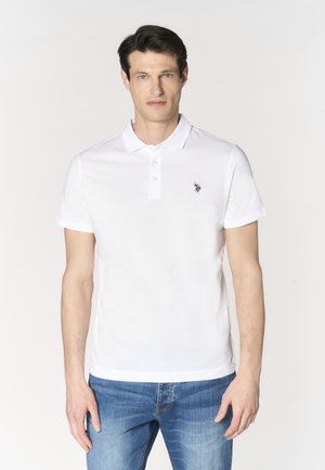 Uomo con capelli corti e scuri che indossa una polo bianca a maniche corte con un piccolo logo sul petto e jeans blu, in piedi contro uno sfondo semplice.