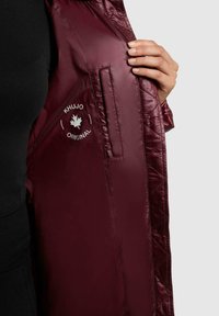 Bordeaux gewatteerde jas interieur met een gladde textuur, voorzien van een zak en een logo met een esdoornblad en de tekst "KHUJO ORIGINAL".