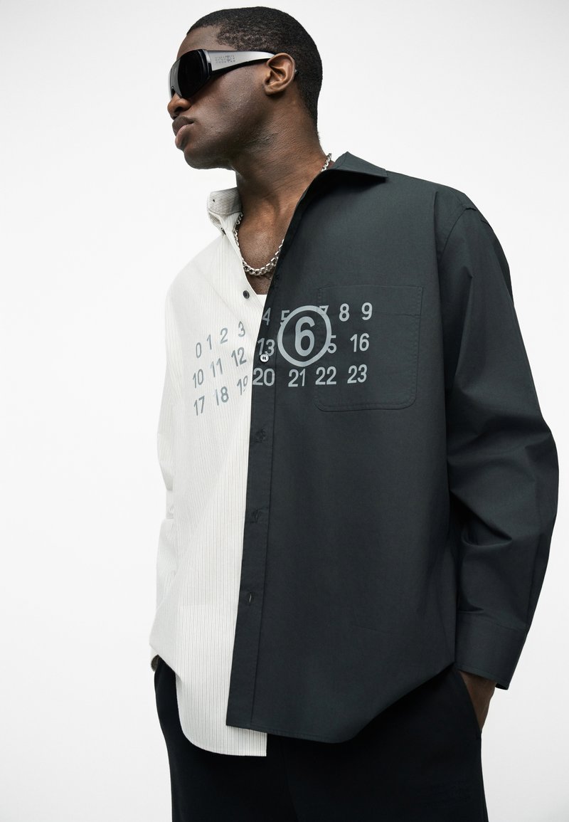 Mann trägt ein halbweißes, halb schwarzes Hemd mit Knöpfen, auf jeder Seite Zahlen gedruckt, schwarze Sonnenbrille und eine silberne Kettenkette.